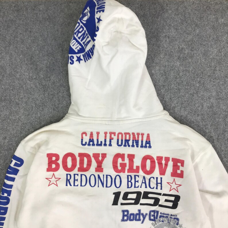 以下が含まれることがあります： 「CALIFORNIA BODY GLOVE REDONDO BEACH 1953」と書かれた青と赤の文字が入った白いパーカー。フードには「CALIFORNIA BODY GLOVE」と書かれた青い文字が入っています。「CALIFORNIA」の文字も左側に縦にプリントされています。