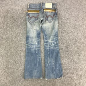 K&ouml;nnte beinhalten: Hellblaue Jeans mit ausgestelltem Bein. Merkmale sind goldfarbene Rei&szlig;verschlusstaschen auf der R&uuml;ckseite, rote N&auml;hte und eine Used-Waschung. Die Jeans haben ein verwaschenes Aussehen mit wei&szlig;en Distressed-Linien.