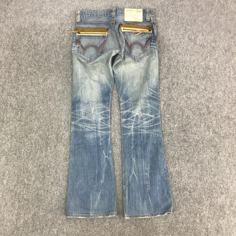 K&ouml;nnte beinhalten: Hellblaue Jeans mit ausgestelltem Bein. Merkmale sind goldfarbene Rei&szlig;verschlusstaschen auf der R&uuml;ckseite, rote N&auml;hte und eine Used-Waschung. Die Jeans haben ein verwaschenes Aussehen mit wei&szlig;en Distressed-Linien.