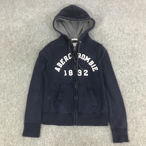 May include: A navy blue zip-up hoodie featuring a grey-lined hood with drawstrings. The front displays white text reading ABERCROMBIE and the number 1892. The garment includes two front pockets, ribbed cuffs, and a ribbed hem for a structured fit.
