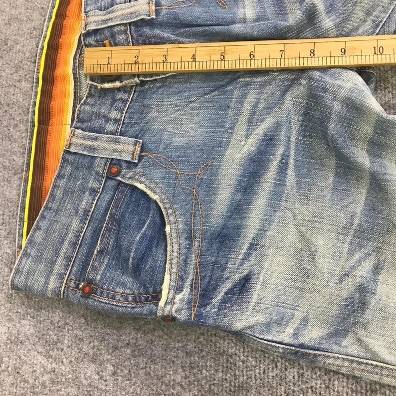 K&ouml;nnte beinhalten: Nahaufnahme einer blauen Jeans mit einem farbenfrohen Seitenstreifen in Gelb, Orange und Braun. Die Jeans hat ein klassisches F&uuml;nf-Taschen-Design und ist gefaltet. Ein Holzlineal liegt auf der Jeans.