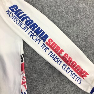 以下が含まれることがあります： 袖に青と赤の文字が入った白い長袖シャツ。「CALIFORNIA SURF GROOVE PROTECTION FROM THE HARSH ELEMENTS」と書かれています。袖口には青いトリムが付いています。