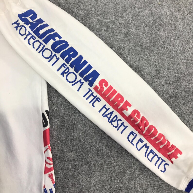 以下が含まれることがあります： 袖に青と赤の文字が入った白い長袖シャツ。「CALIFORNIA SURF GROOVE PROTECTION FROM THE HARSH ELEMENTS」と書かれています。袖口には青いトリムが付いています。