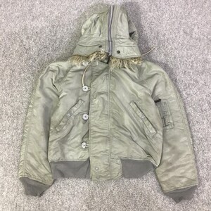 Könnte beinhalten: Eine olivgrüne Bomberjacke mit Kapuze und Kunstfellbesatz. Die Jacke hat einen Reißverschluss, Knöpfe und gerippte Bündchen und Saum. Sie hat zwei Vordertaschen und eine Utility-Tasche am linken Ärmel.