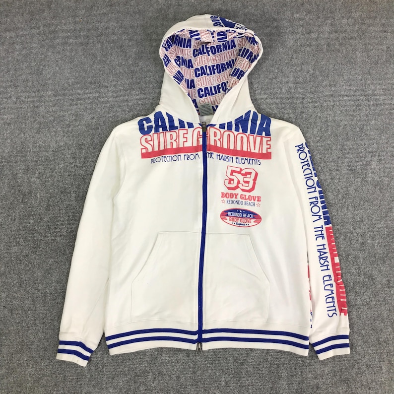 以下が含まれることがあります： 白のジップアップパーカー。青と赤のアクセントが特徴です。「CALIFORNIA SURF GROOVE」と「BODY GLOVE REDONDO BEACH」の文字と数字「53」がプリントされています。フードの裏地には「CALIFORNIA」の繰り返しパターンがあります。