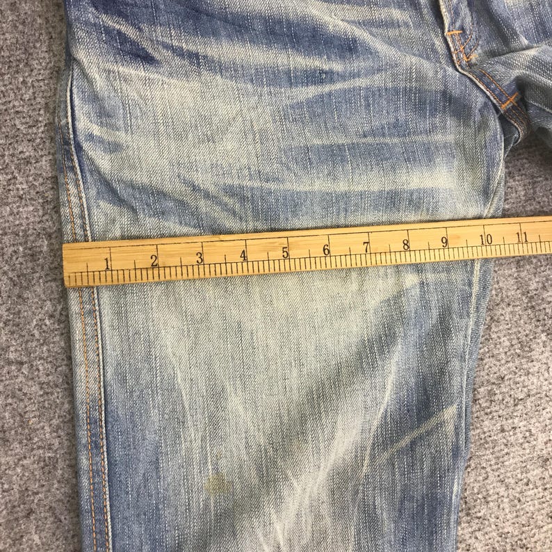 K&ouml;nnte beinhalten: Nahaufnahme einer hellblauen Jeans mit einem Holzlineal auf dem Stoff. Das Lineal zeigt Messungen in Zentimetern an, die die Abmessungen der Jeans angeben. Der Denim hat ein verwaschenes, abgenutztes Aussehen.