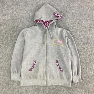 May include: A gray zip-up hoodie featuring a Hello Kitty design. The hoodie has a hood with pink leopard print lining and a matching bow. The pockets also have leopard print accents. The Hello Kitty graphic is on the front.