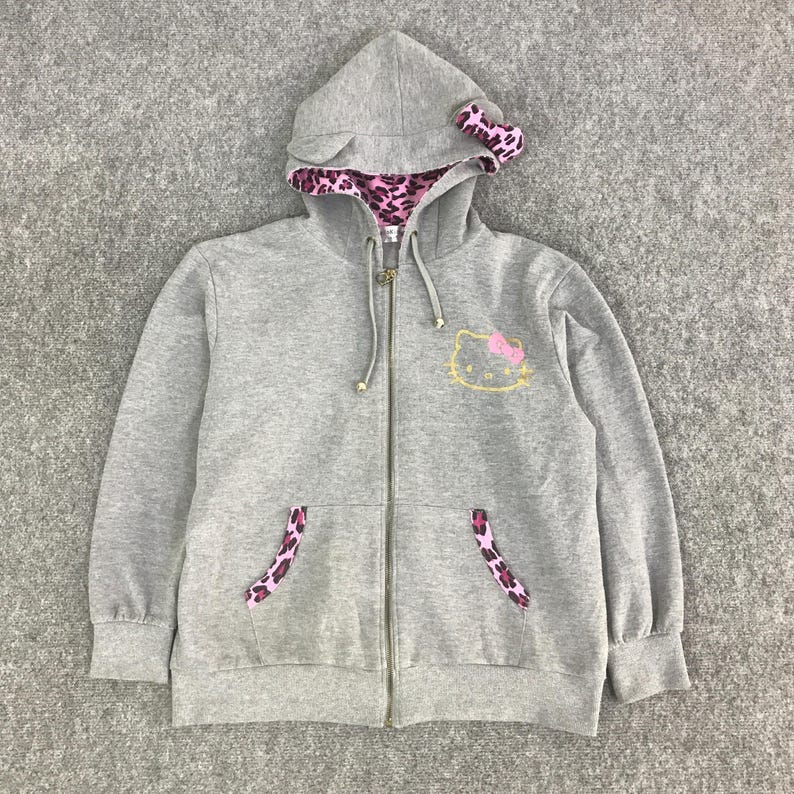 May include: A gray zip-up hoodie featuring a Hello Kitty design. The hoodie has a hood with pink leopard print lining and a matching bow. The pockets also have leopard print accents. The Hello Kitty graphic is on the front.