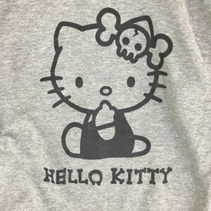 Może przedstawiać: Szara bluza z czarną grafiką Hello Kitty z kokardą w kształcie czaszki i napisem "HELLO KITTY" poniżej. Projekt jest umieszczony na przodzie bluzy.