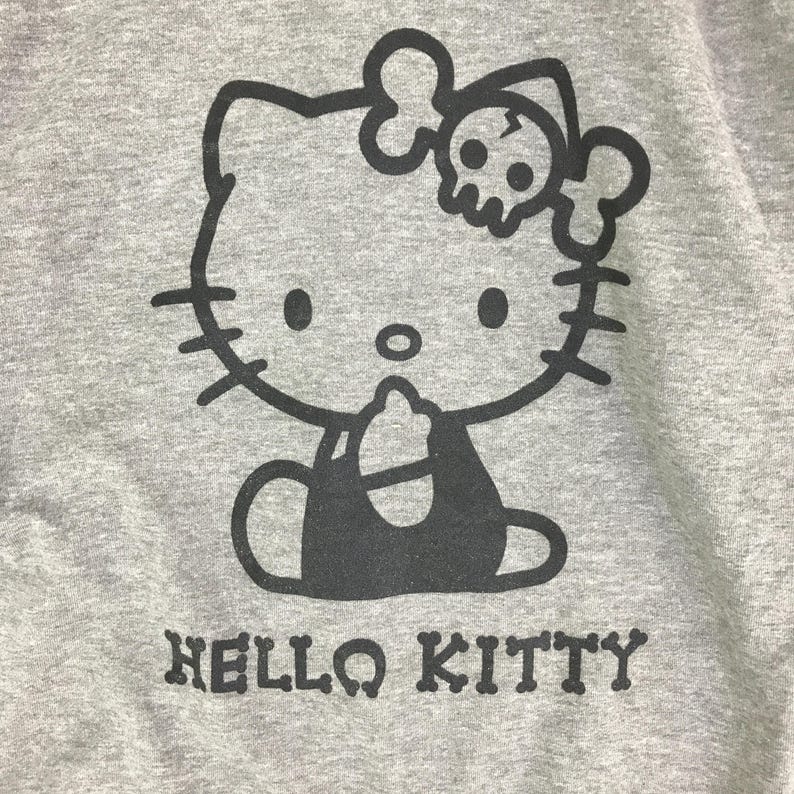 Może przedstawiać: Szara bluza z czarną grafiką Hello Kitty z kokardą w kształcie czaszki i napisem "HELLO KITTY" poniżej. Projekt jest umieszczony na przodzie bluzy.