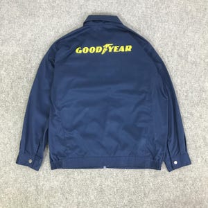 Könnte beinhalten: Marineblaue Jacke mit gelbem "GOOD YEAR"-Schriftzug auf dem Rücken. Die Jacke hat eine Knopfleiste, geknöpfte Manschetten und einen gerippten Saum. Hergestellt aus einem glatten, strapazierfähigen Stoff.