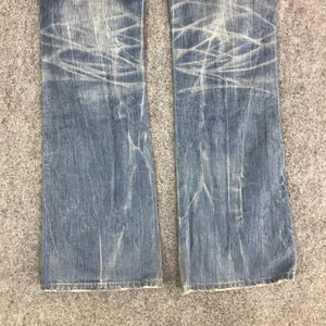 K&ouml;nnte beinhalten: Hellblaue Jeans aus Denim mit wei&szlig;en, abgenutzten Markierungen. Die Jeans haben eine verblasste Waschung und einen leichten Schlag am unteren Saum. Der Stoff weist Abnutzungserscheinungen auf, was ihnen einen Vintage-Look verleiht. Die Jeans liegen flach auf einer grauen Oberfl&auml;che.