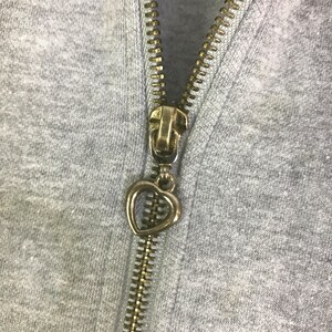 May include: Close-up of a gray hoodie zipper with a gold-colored metal zipper pull. The zipper pull is in the shape of a heart. The zipper teeth are also gold-colored.