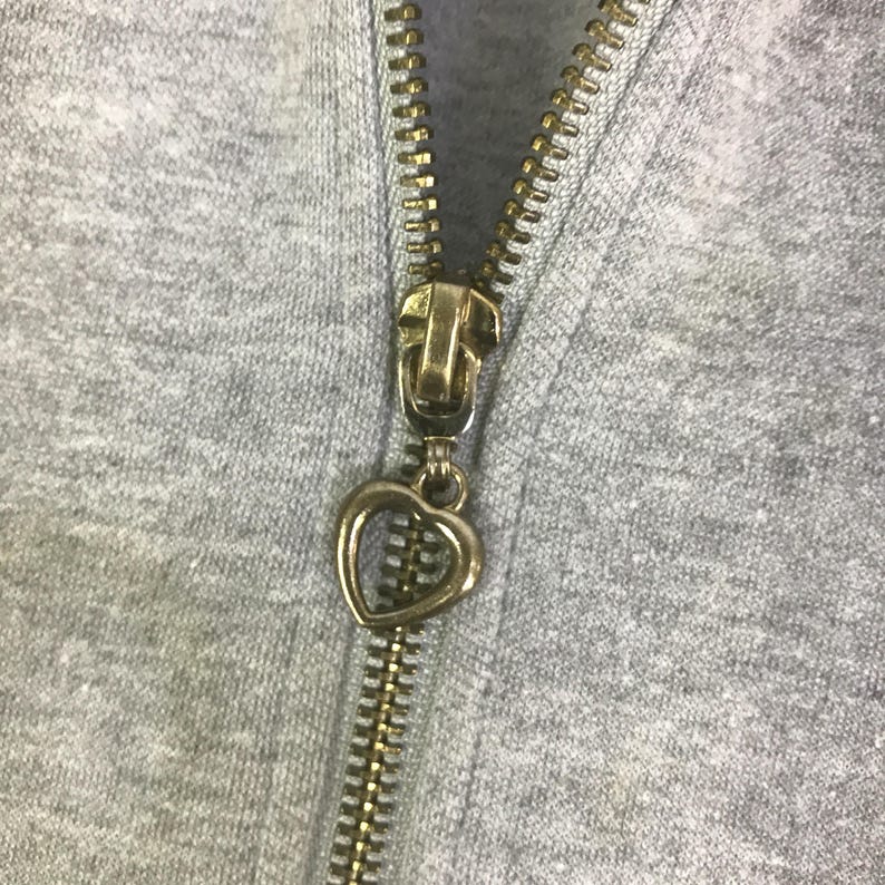 May include: Close-up of a gray hoodie zipper with a gold-colored metal zipper pull. The zipper pull is in the shape of a heart. The zipper teeth are also gold-colored.