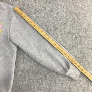 May include: A gray sweatshirt sleeve with a pink and gold design. A wooden ruler is placed diagonally across the sleeve, measuring up to 22 inches. The sleeve has a ribbed cuff and a small stain.