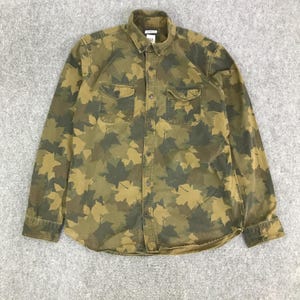 May include: A long-sleeved button-down shirt featuring a camouflage design of green and tan maple leaves. The shirt has two chest pockets and a collar. The shirt is made of a durable fabric.