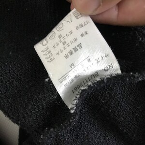 May include: White tag with black text detailing the fabric composition of a black garment. The tag reads: "Body: Polyester 73%, Cotton 27%, Rib: Polyester 80%, Cotton 20%". The tag also includes size information: "Size: M, Bust: 79-87 cm, Length: 154-162 cm".