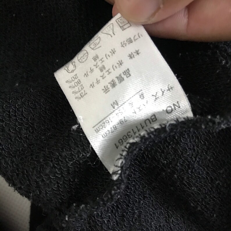 May include: White tag with black text detailing the fabric composition of a black garment. The tag reads: "Body: Polyester 73%, Cotton 27%, Rib: Polyester 80%, Cotton 20%". The tag also includes size information: "Size: M, Bust: 79-87 cm, Length: 154-162 cm".