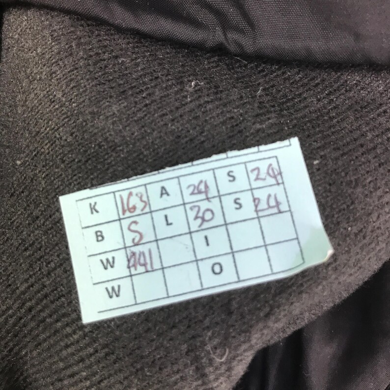 May include: A dark brown, textured fabric garment featuring a light blue rectangular tag with a grid pattern. The tag contains handwritten black ink notations including K 163, A 24, S 24, B S, L 30, S 24, and W 41, providing specific garment measurements.