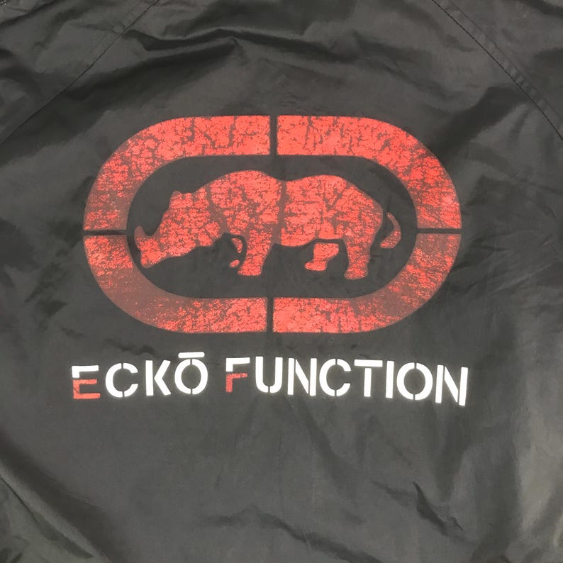 May include: A black jacket featuring a large red graphic of a rhinoceros inside an oval frame with a distressed texture. Below the graphic, the text ECKO FUNCTION is printed in white, with the letters E and F highlighted in a distressed red color.