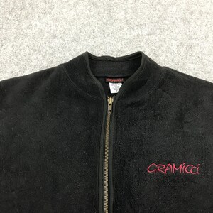 以下が含まれることがあります： 黒色のフリースジップアップジャケット。胸元に赤色で「Gramicci」のブランド名が刺繍されています。ジャケットはハイカラーで、フルレングスのジッパーが付いています。生地は柔らかく、質感があります。