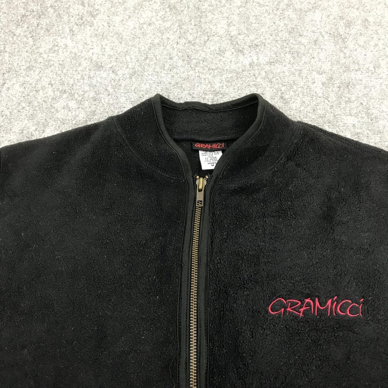以下が含まれることがあります： 黒色のフリースジップアップジャケット。胸元に赤色で「Gramicci」のブランド名が刺繍されています。ジャケットはハイカラーで、フルレングスのジッパーが付いています。生地は柔らかく、質感があります。