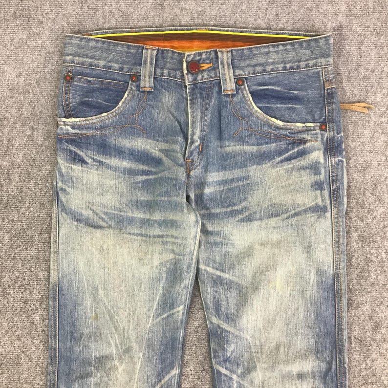 K&ouml;nnte beinhalten: Verblasste blaue Jeans aus Denim mit einem Used-Look. Die Jeans haben ein klassisches F&uuml;nf-Taschen-Design, einen Knopf- und Rei&szlig;verschluss und orangefarbene Nahtdetails. Der Bund hat ein gelb-oranges Innenfutter.