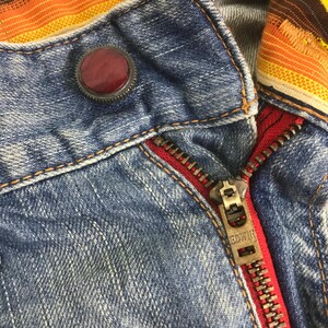 K&ouml;nnte beinhalten: Nahaufnahme einer Jeansjacke mit rotem Rei&szlig;verschluss und dekorativem Knopf. Der Rei&szlig;verschluss ist aus Metall und der Knopf hat eine dunkelrote Farbe. Die Jacke hat ein buntes Streifenmuster am Kragen.