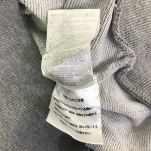 Może przedstawiać: Zbliżenie szarej bluzy z dołączoną białą metką. Etykieta zawiera japoński tekst i instrukcje pielęgnacji. Bluza wykonana jest z miękkiej, teksturowanej tkaniny.