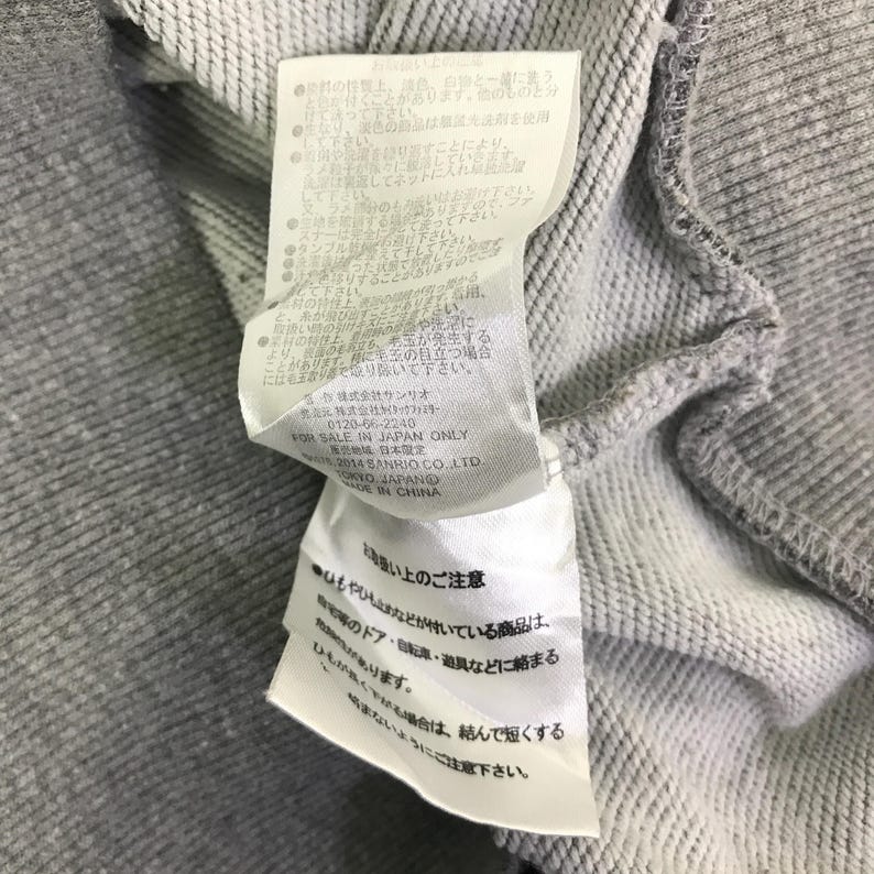 Może przedstawiać: Zbliżenie szarej bluzy z dołączoną białą metką. Etykieta zawiera japoński tekst i instrukcje pielęgnacji. Bluza wykonana jest z miękkiej, teksturowanej tkaniny.