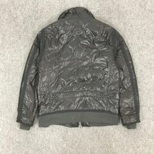 Może przedstawiać: Czarna pikowana kurtka ze st&oacute;jką i prążkowanym dołem. Kurtka posiada wz&oacute;r w romby i zapięcie na zamek błyskawiczny. Rękawy mają czarne paski po bokach. Kurtka wykonana jest z błyszczącego materiału.