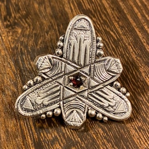 May include: A silver pendant with a geometric design and a red gemstone in the center. The pendant is shaped like a star and has a beaded border.