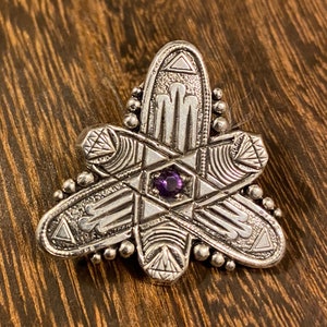May include: A silver pendant with a geometric design and a purple gemstone in the center.
