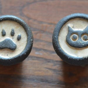 Könnte beinhalten: Zwei kreisförmige Keramikknöpfe mit einem dunkel anthrazitfarbenen Rand und einer cremefarbenen Mitte. Ein Knopf zeigt ein erhabenes schwarzes Pfotenabdruck-Design, während der andere Knopf ein schwarzes Katzenkopf-Symbol mit zwei runden Augen zeigt.