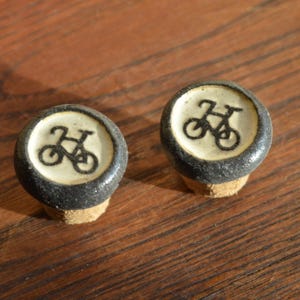 Puede incluir: Dos tapones de botella de cerámica en blanco y negro con un diseño de bicicleta. Los tapones están respaldados por corcho.