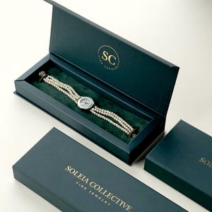 May include: A teal jewelry box with gold lettering reading "SOLEIA COLLECTIVE FINE JEWELRY" and a watch with a pearl band. The watch is displayed in an open box with a green velvet interior. The box has the initials "SC" in gold.
