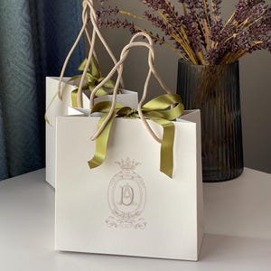 May include: Two white gift bags with beige rope handles and olive green ribbon bows. Each bag features a light brown emblem with the letters "AD" and a crown. A vase of dried lavender is in the background.