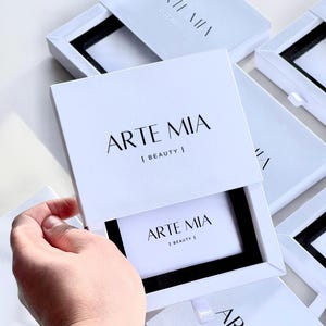 Pode incluir: Caixas brancas com o texto "ARTE MIA | BEAUTY |" em preto. Uma caixa está aberta, revelando um interior preto e um cartão com o mesmo texto. Várias outras caixas são visíveis, algumas abertas, outras fechadas.