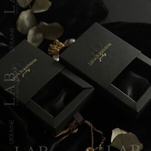 May include: Two black jewelry boxes with gold lettering that reads "Lena Riabinnina Jewelry". The boxes are open and lined with black velvet. A gold chain necklace is lying on a black surface.