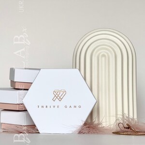 May include: White hexagonal box with the text "THRIVE GANG" and a gold heart with a diamond inside. The box is sitting on a stack of smaller white boxes with pink lids. A white ceramic arch with a ribbed design is behind the box. A gold bracelet is lying on a pink feather.