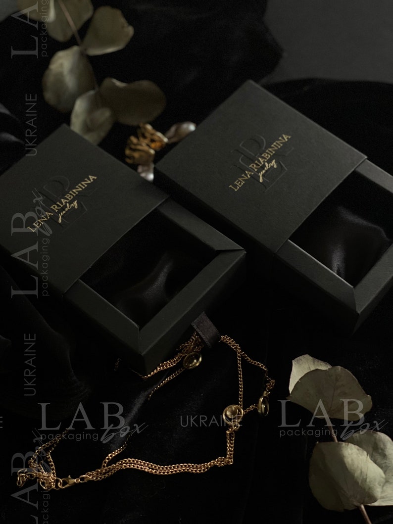 May include: Two black jewelry boxes with gold lettering that reads "Lena Riabinnina Jewelry". The boxes are open and lined with black velvet. A gold chain necklace is lying on a black surface.