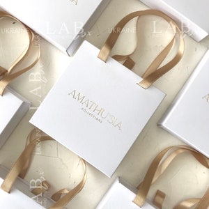 K&ouml;nnte beinhalten: Wei&szlig;e Geschenkbox mit goldener Schleife und dem Aufdruck "AMATHUSIA COLLECTIONS" auf der Vorderseite.