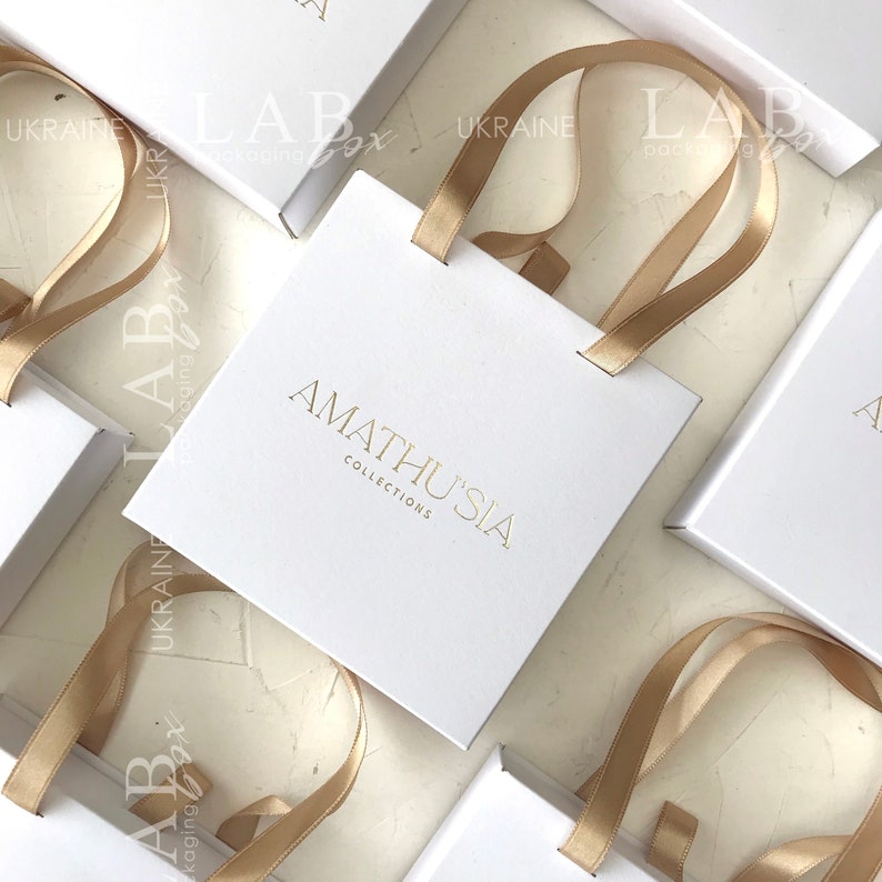 K&ouml;nnte beinhalten: Wei&szlig;e Geschenkbox mit goldener Schleife und dem Aufdruck "AMATHUSIA COLLECTIONS" auf der Vorderseite.