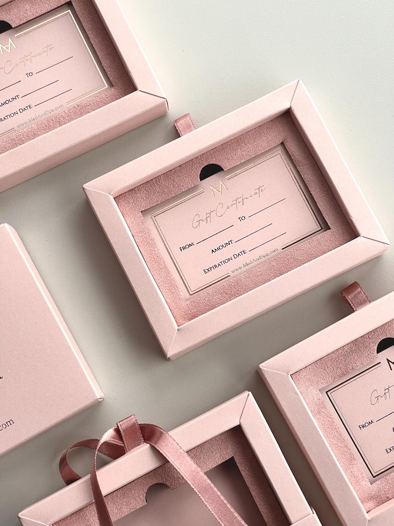 May include: Pink gift certificate boxes with a velvet interior and a ribbon pull. Each box contains a gift certificate with the text "Gift Certificate" and fields for "From", "To", "Amount", and "Expiration Date". The brand logo "M" is visible.