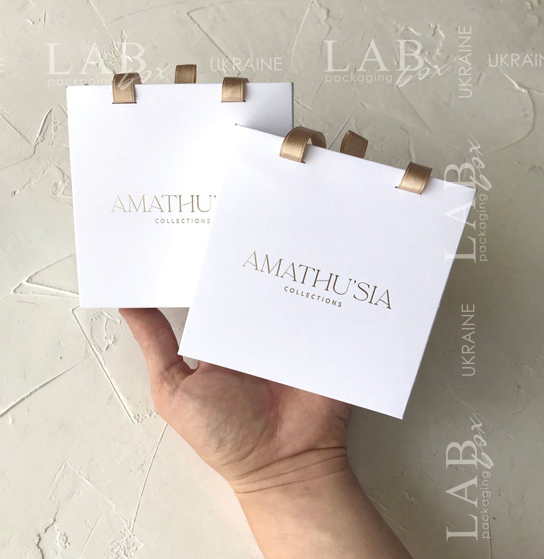 K&ouml;nnte beinhalten: Zwei wei&szlig;e Geschenkboxen mit goldener Schleife und dem aufgedruckten Text "AMATHU' COLLECTIONS" und "AMATHUSIA COLLECTIONS".