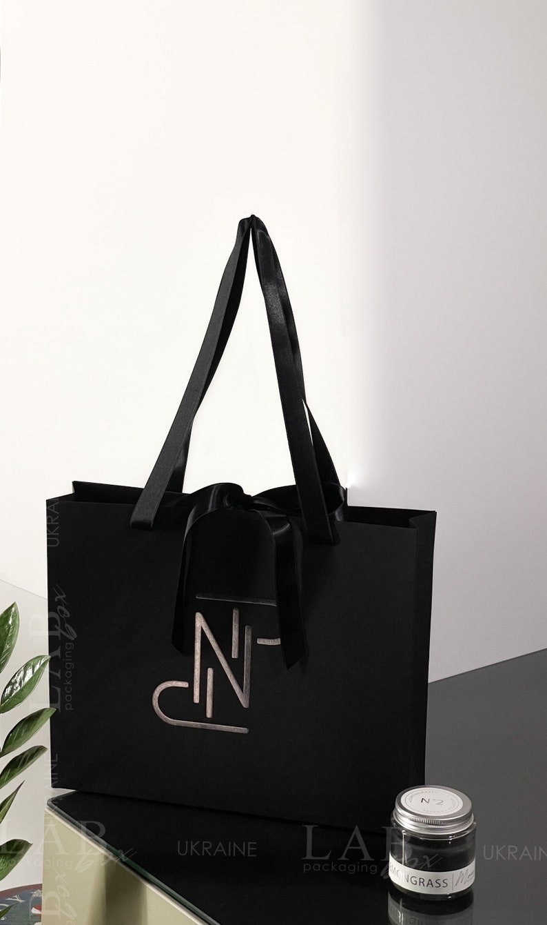 Puede incluir: Una bolsa de regalo negra con un logotipo plateado que dice "N-N" y una cinta de sat&eacute;n negra. La bolsa est&aacute; sobre una mesa con un peque&ntilde;o frasco etiquetado como "N 2" y "Lemongrass".