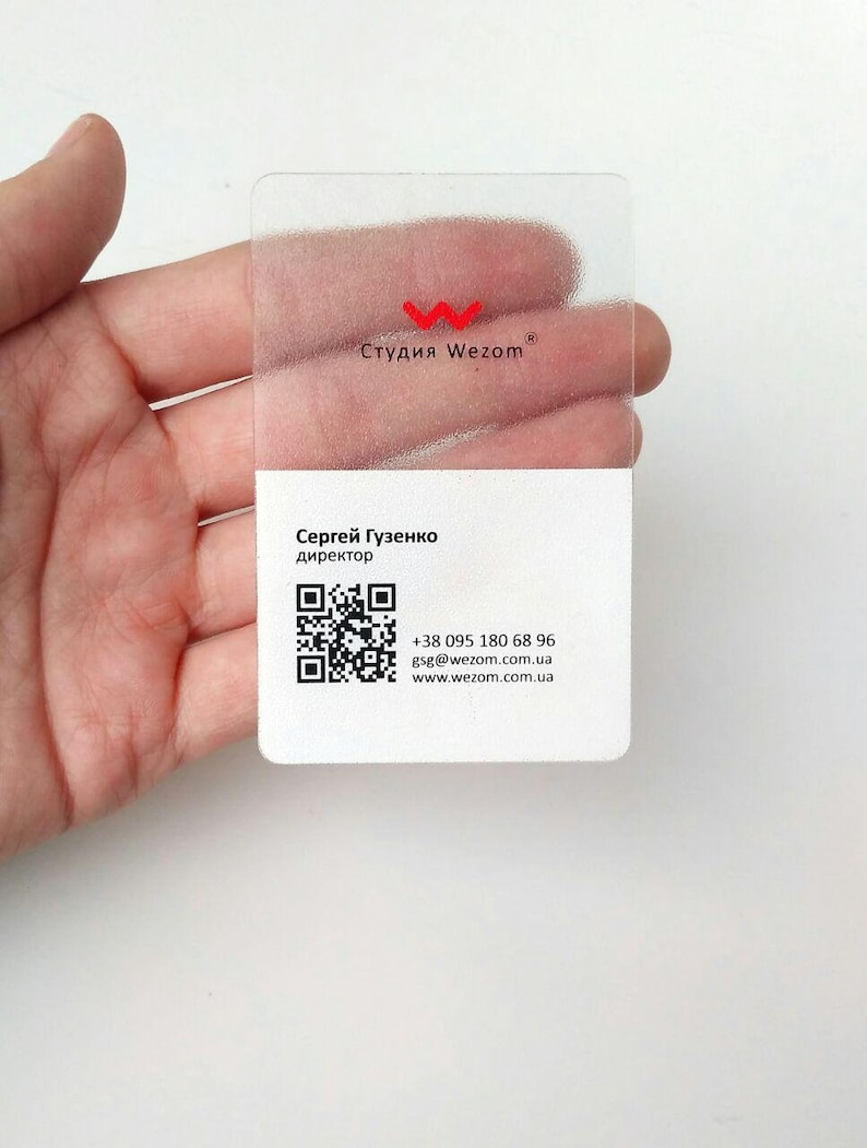 Op de afbeelding: Een transparante plastic visitekaartje met een rood "W" logo en de tekst "Студия Wezom" in Cyrillisch schrift. De kaart bevat ook de naam "Сергей Гузенко", de titel "директор", een QR-code, een telefoonnummer, een e-mailadres en een websiteadres.