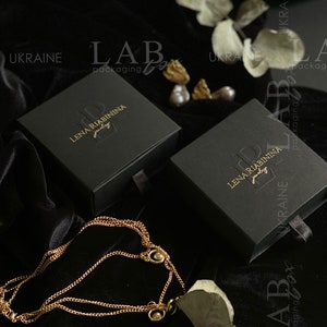 May include: Two black jewelry boxes with gold lettering. The boxes are labeled "Lena Riabinina Jewelry" and "Lab Packaging Box". The boxes are on a black velvet surface with a gold chain necklace.