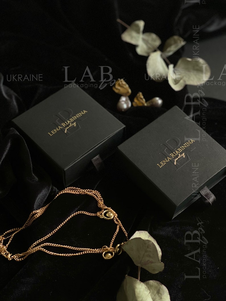 May include: Two black jewelry boxes with gold lettering. The boxes are labeled "Lena Riabinina Jewelry" and "Lab Packaging Box". The boxes are on a black velvet surface with a gold chain necklace.