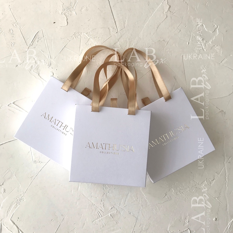 K&ouml;nnte beinhalten: Drei wei&szlig;e Geschenkt&uuml;ten mit goldenen Henkeln und dem Text "AMATHUSIA COLLECTIONS" in Gold auf der Vorderseite.